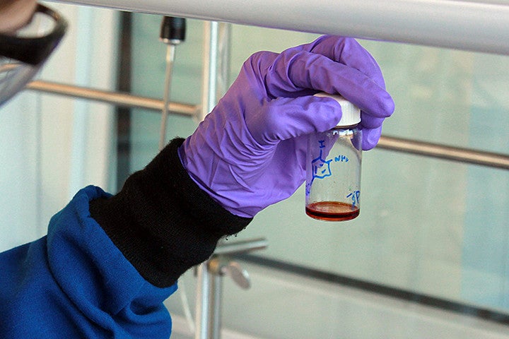 A solution in a container being held in a hand wearing a purple glove