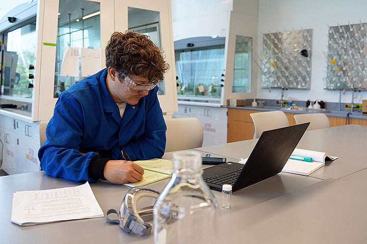 A person in a blue lab coat does work on a laptop in a lab