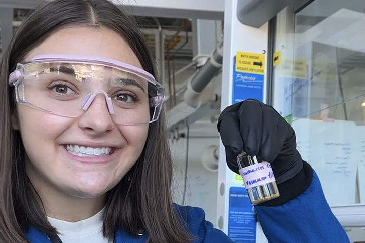 A person holds up a solution in a vial while smiling at the camera