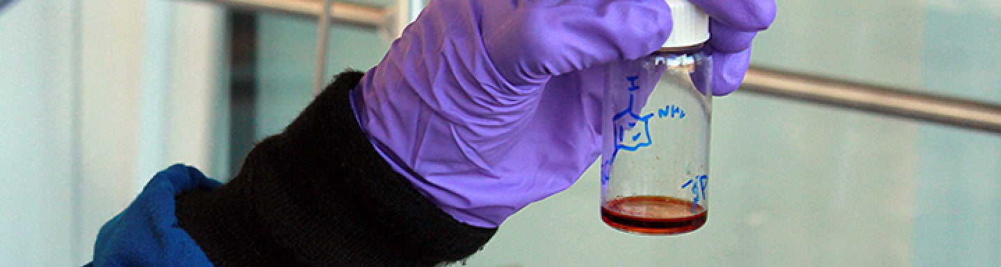 A solution in a container being held in a hand wearing a purple glove
