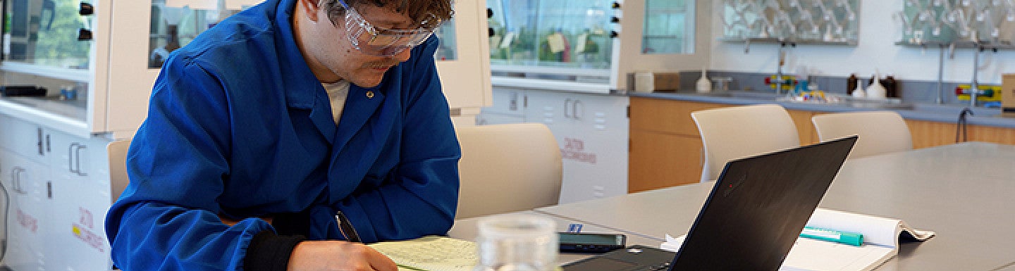 A person in a blue lab coat does work on a laptop in a lab